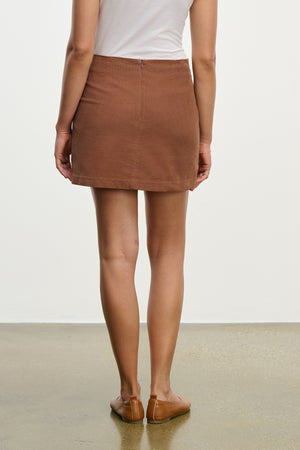 A person wearing a white sleeveless top, the Velvet by Graham & Spencer LIZA CORDUROY SKIRT, and brown flat shoes stands on a polished floor facing away from the camera.