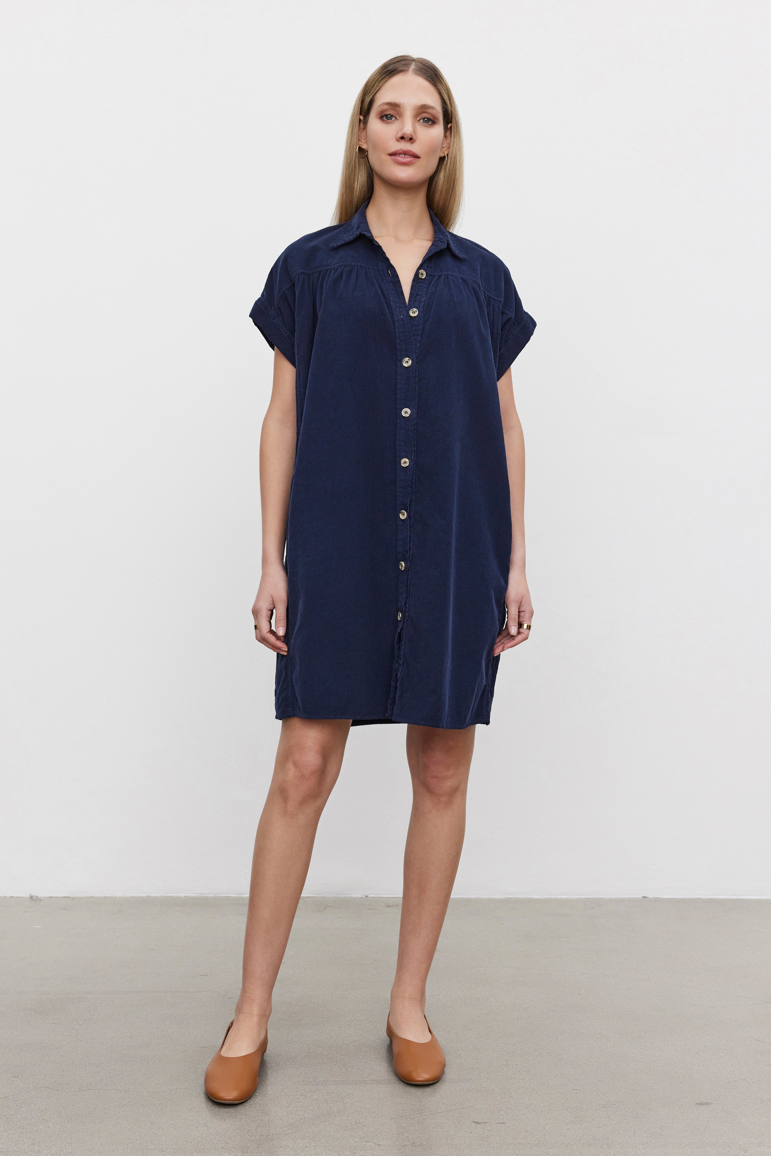   A woman is standing, wearing the MACIE CORDUROY DRESS by Velvet by Graham & Spencer, a short-sleeve navy blue button-down dress with a classic collar. She pairs it with brown shoes and faces forward against a plain white background. 