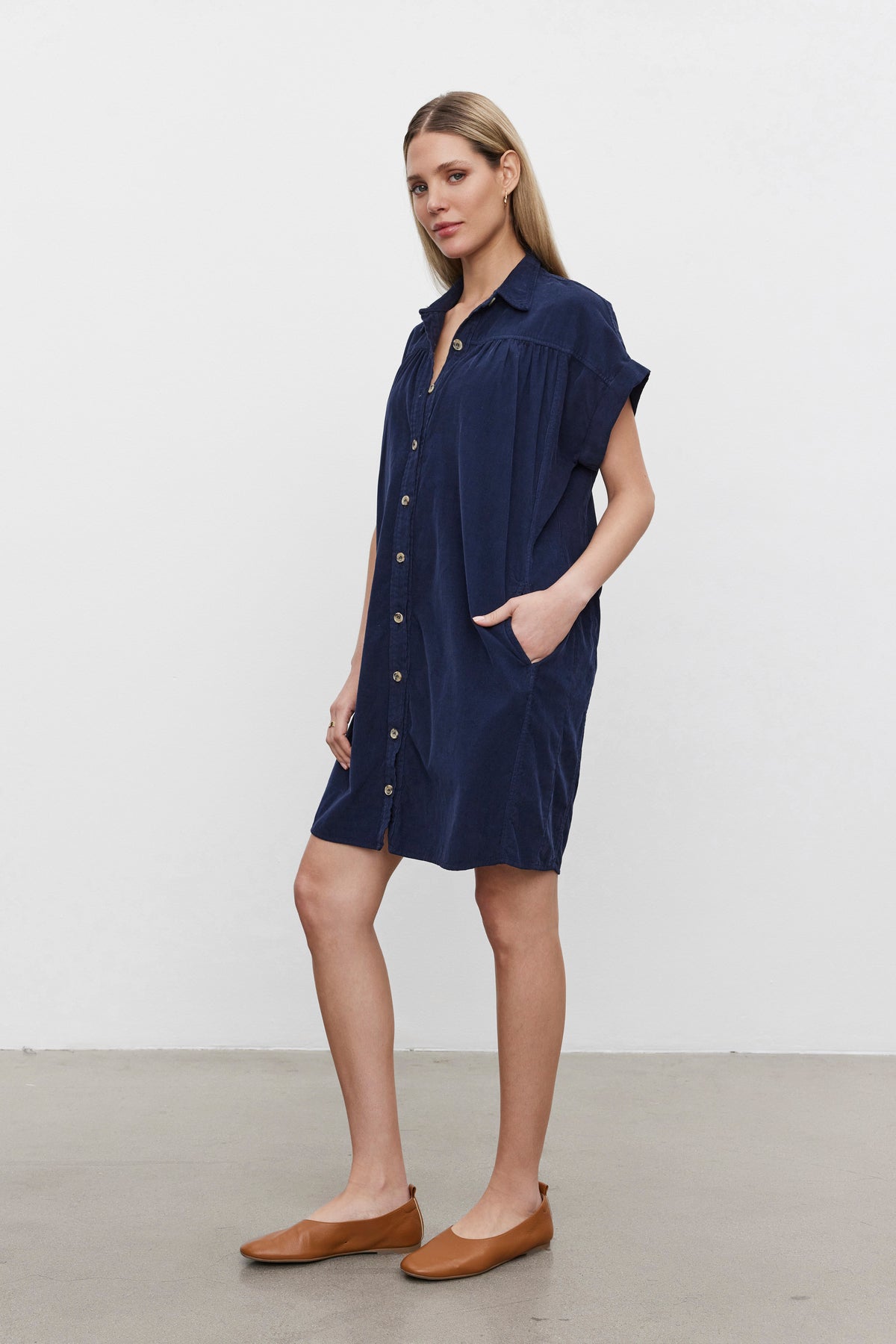   A woman stands against a plain background wearing the MACIE CORDUROY DRESS by Velvet by Graham & Spencer, which features short sleeves, a classic collar, and is paired with brown flat shoes. 