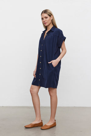 A woman stands against a plain background wearing the MACIE CORDUROY DRESS by Velvet by Graham & Spencer, which features short sleeves, a classic collar, and is paired with brown flat shoes.