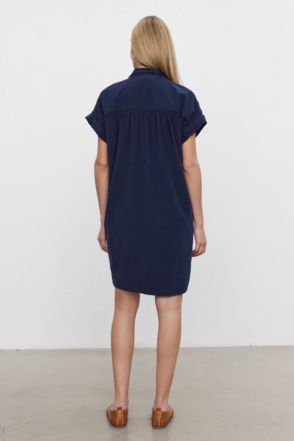   A woman with blonde hair, standing and facing away, wears the MACIE CORDUROY DRESS by Velvet by Graham & Spencer in navy blue, featuring short sleeves and a classic collar, paired with brown shoes against a plain background. 