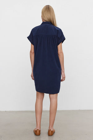 A woman with blonde hair, standing and facing away, wears the MACIE CORDUROY DRESS by Velvet by Graham & Spencer in navy blue, featuring short sleeves and a classic collar, paired with brown shoes against a plain background.