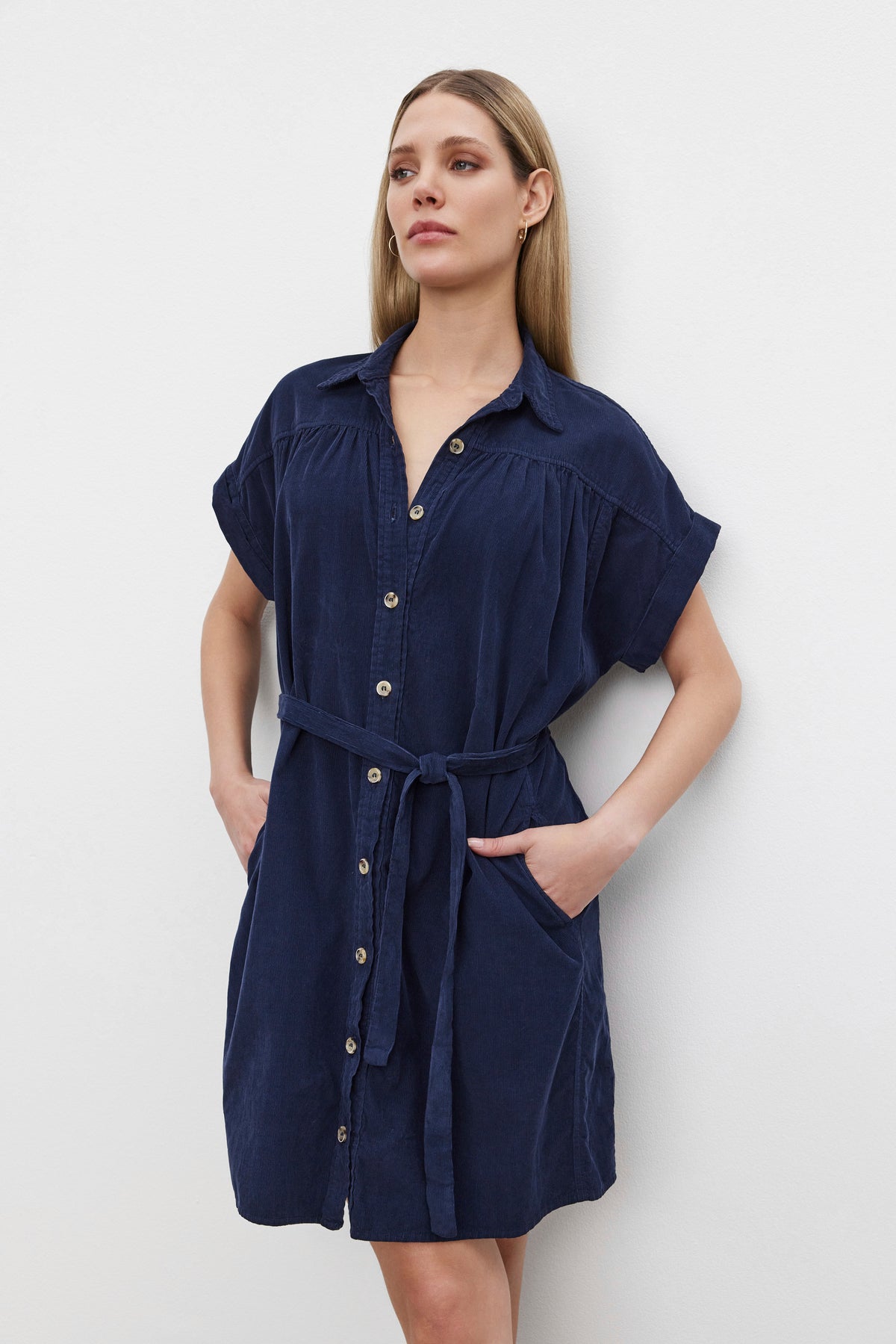   A woman with long hair wearing the MACIE CORDUROY DRESS, a navy blue button-down dress with a classic collar and a detachable belt by Velvet by Graham & Spencer, stands against a plain white background. 