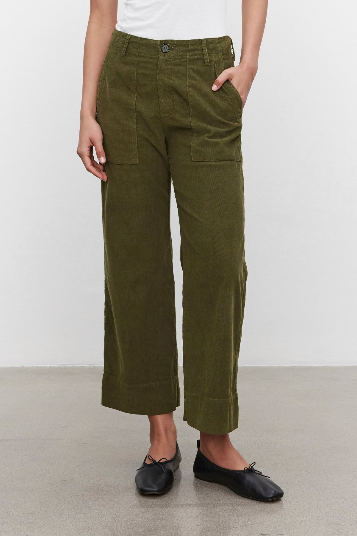   A person is wearing a white top, VERA CORDUROY PANT in olive green by Velvet by Graham & Spencer, and black flats. They have one hand in a pocket and are standing against a plain white background. 