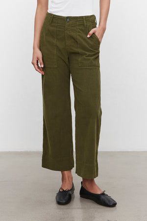 A person is wearing a white top, VERA CORDUROY PANT in olive green by Velvet by Graham & Spencer, and black flats. They have one hand in a pocket and are standing against a plain white background.