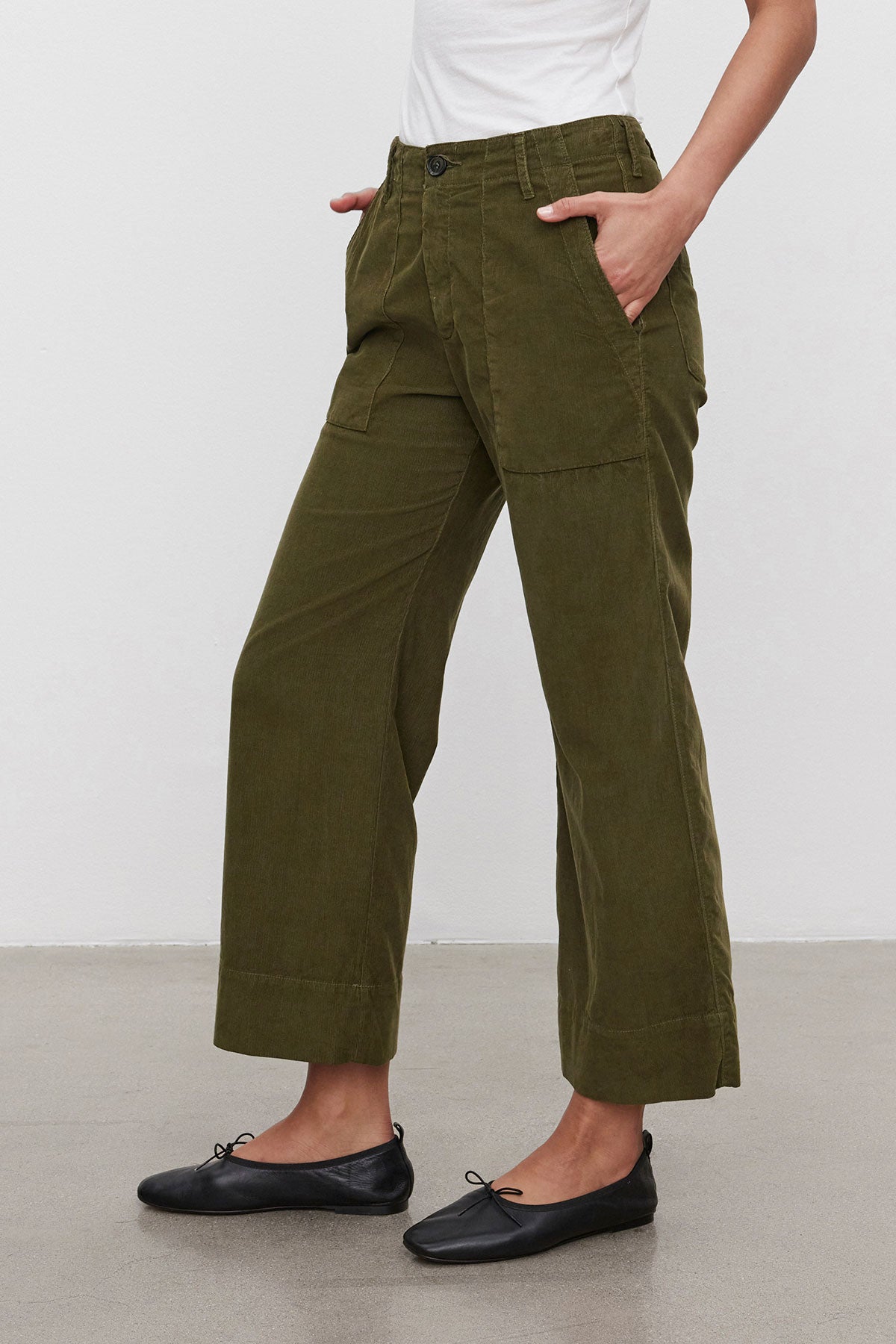   A person is wearing the VERA CORDUROY PANT by Velvet by Graham & Spencer in olive green, featuring wide legs and utility details, with their hands in the pockets. They are also wearing black ballet flats against a plain white background. 