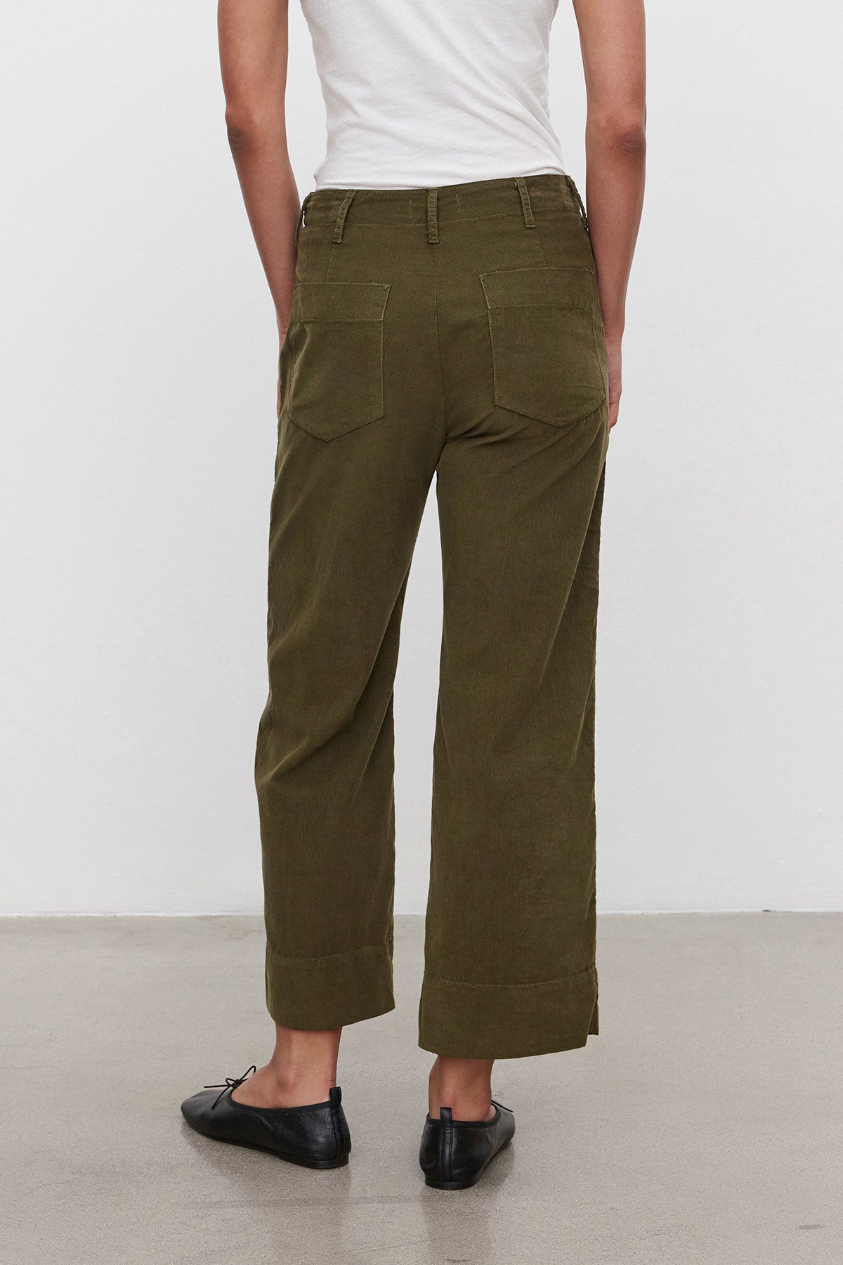   A person stands with their back to the camera, donning a white top, VERA CORDUROY PANT by Velvet by Graham & Spencer—olive green pinwale corduroy wide-leg pants featuring utility details and pockets—and black flats. 