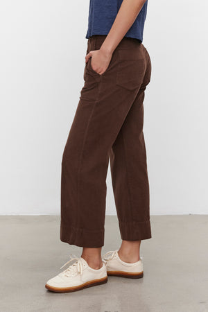 A person wearing the VERA CORDUROY PANT from Velvet by Graham & Spencer, complemented by a blue shirt and cream-colored sneakers, stands on a grey concrete floor with a white wall in the background. The individual is in a relaxed pose with hands in pockets.