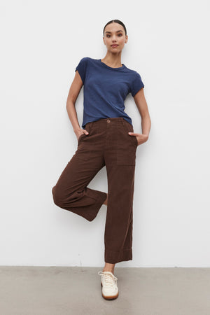 A person stands with hands in pockets, wearing a blue t-shirt, Velvet by Graham & Spencer's VERA CORDUROY PANT in pinwale corduroy cropped wide-leg style, and white sneakers. The backdrop is a plain white wall.