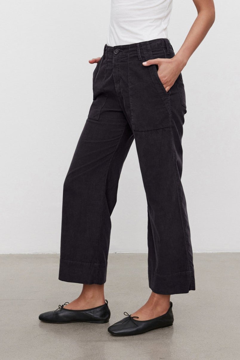 A person is standing against a plain backdrop wearing a white tank top, Velvet by Graham & Spencer's VERA CORDUROY PANT in black, and black flats. Hands are in the pockets, looking down at the ground.-37665605550273