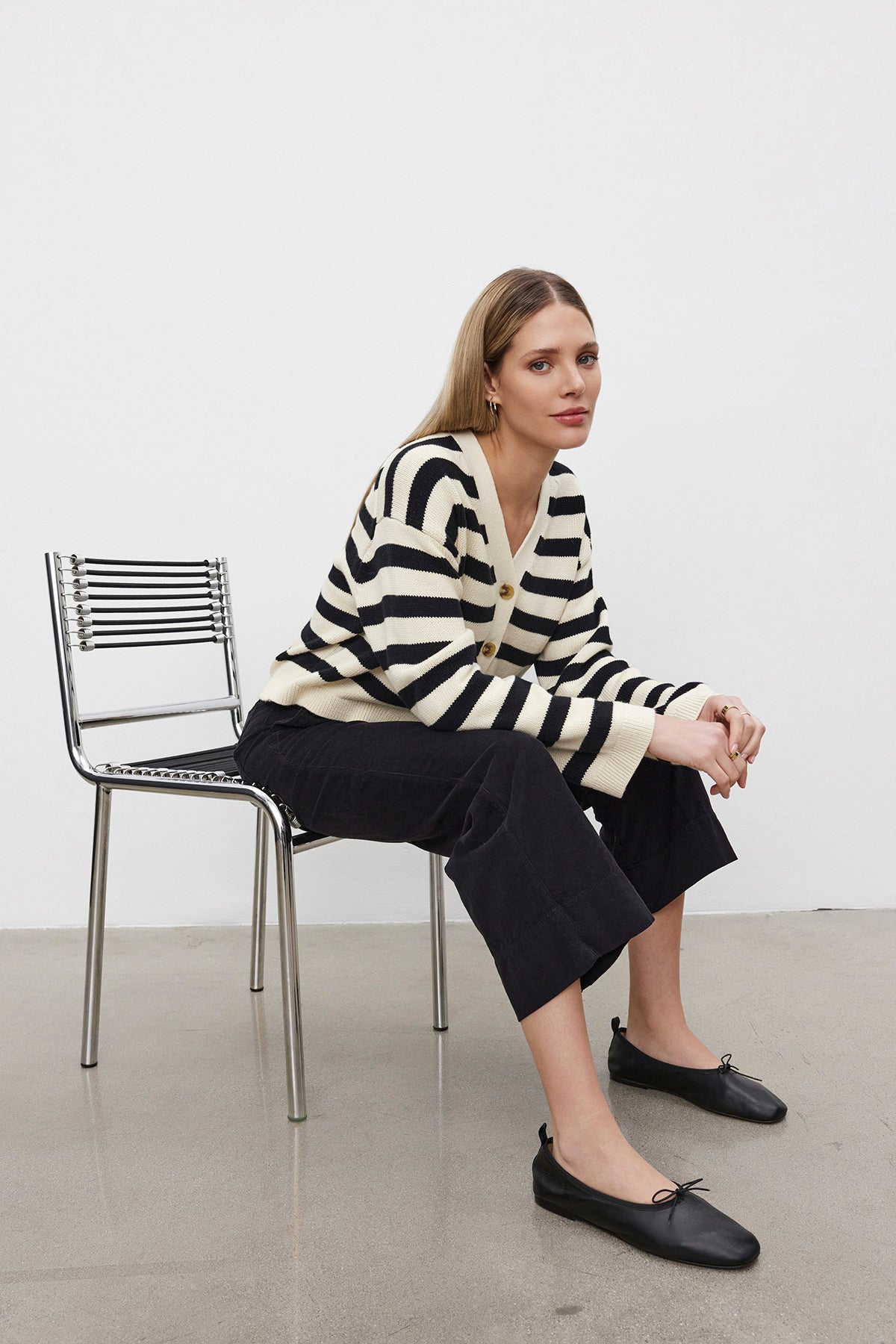   A person wearing a striped sweater, VERA CORDUROY PANT by Velvet by Graham & Spencer, and black shoes sits on a metal chair in front of a white wall. 