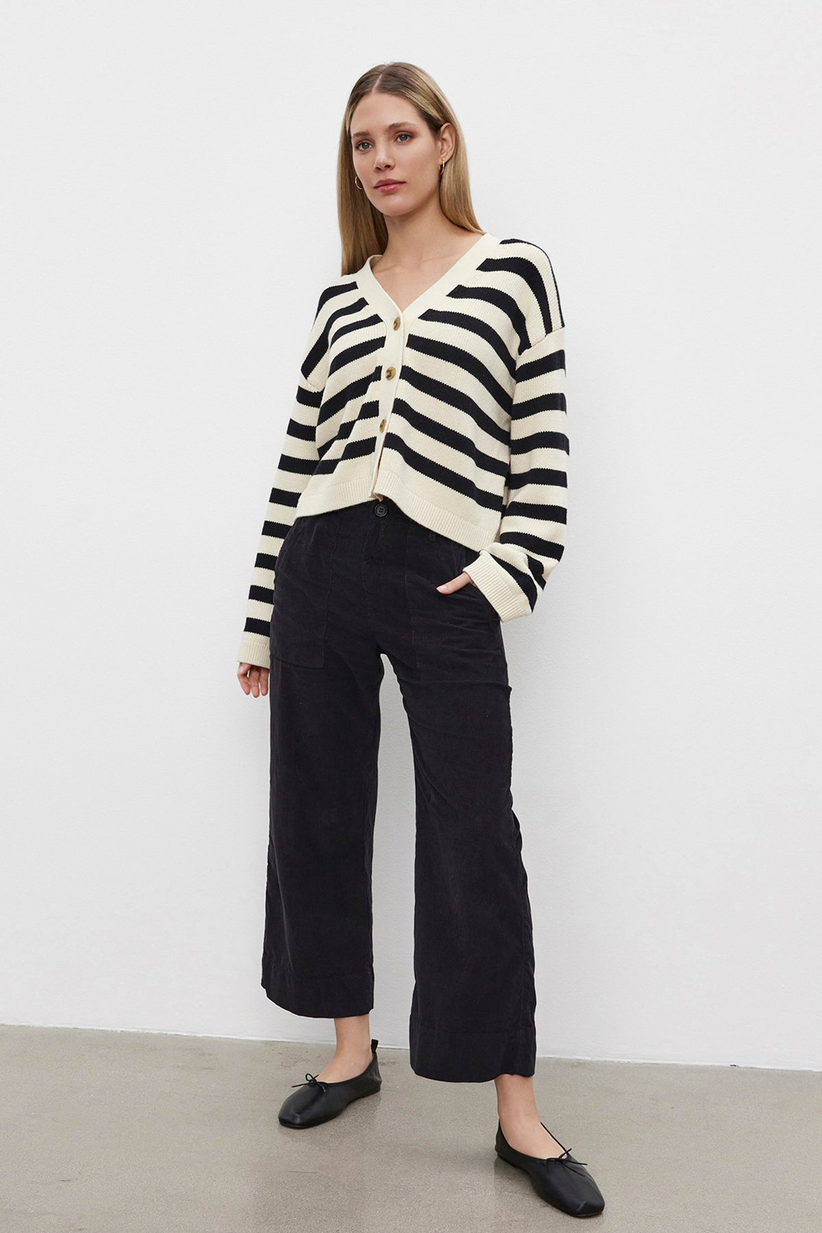   A person stands against a white background, wearing the ELODIE CARDIGAN by Velvet by Graham & Spencer—a cotton cashmere sweater with black and white stripes in a cropped design—paired with black pants and sleek black shoes. 