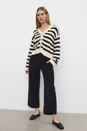 A person stands against a white background, wearing the ELODIE CARDIGAN by Velvet by Graham & Spencer—a cotton cashmere sweater with black and white stripes in a cropped design—paired with black pants and sleek black shoes.