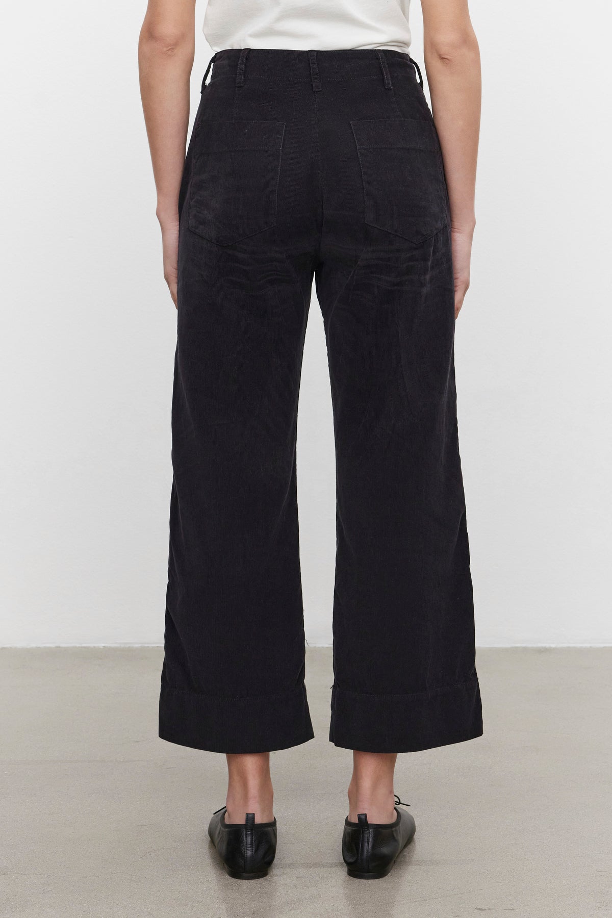   Back view of a person wearing the Velvet by Graham & Spencer VERA CORDUROY PANT in black pinwale corduroy, paired with a white top and black shoes, standing on a grey floor against a plain white background. 