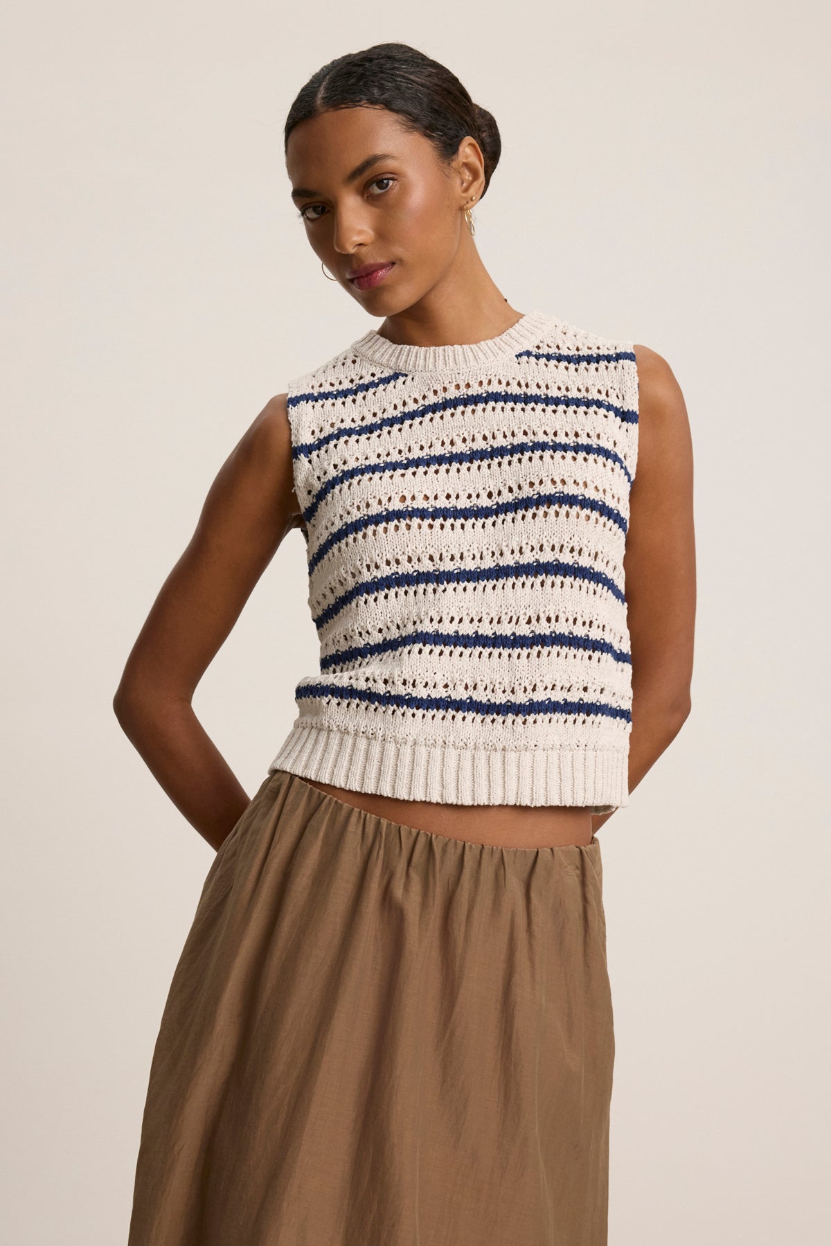 A woman wears the CALISTA SWEATER VEST by Velvet by Graham & Spencer, a sleeveless knit top with blue and cream crochet stripes, styled with a high-waisted light brown skirt against a plain background.