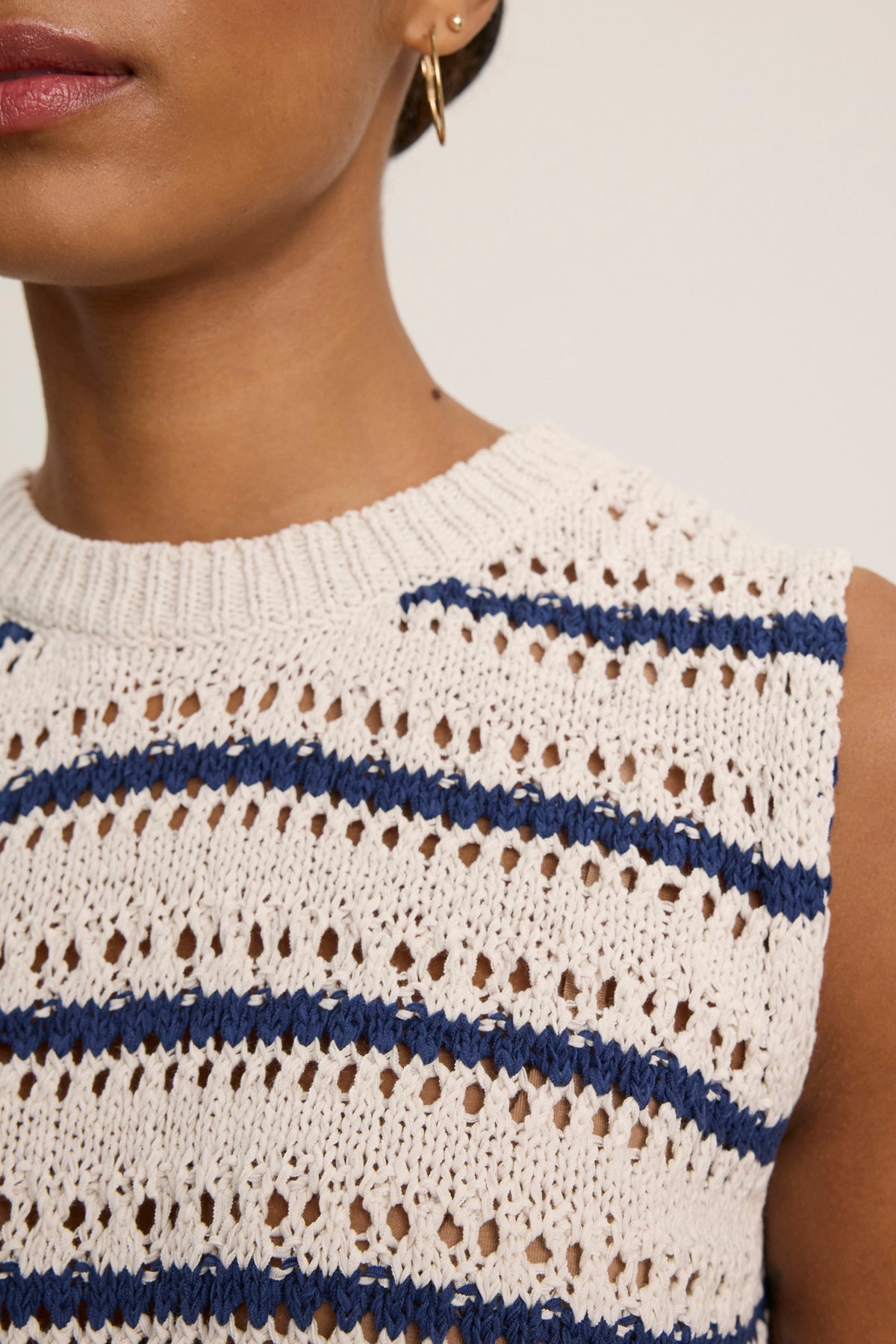 Close-up of a person wearing the CALISTA SWEATER VEST by Velvet by Graham & Spencer—a sleeveless, cream-colored cropped vest with blue stripes and openwork details. Gold hoop earring adds versatility to the chic crochet stitch design.-Hover Image