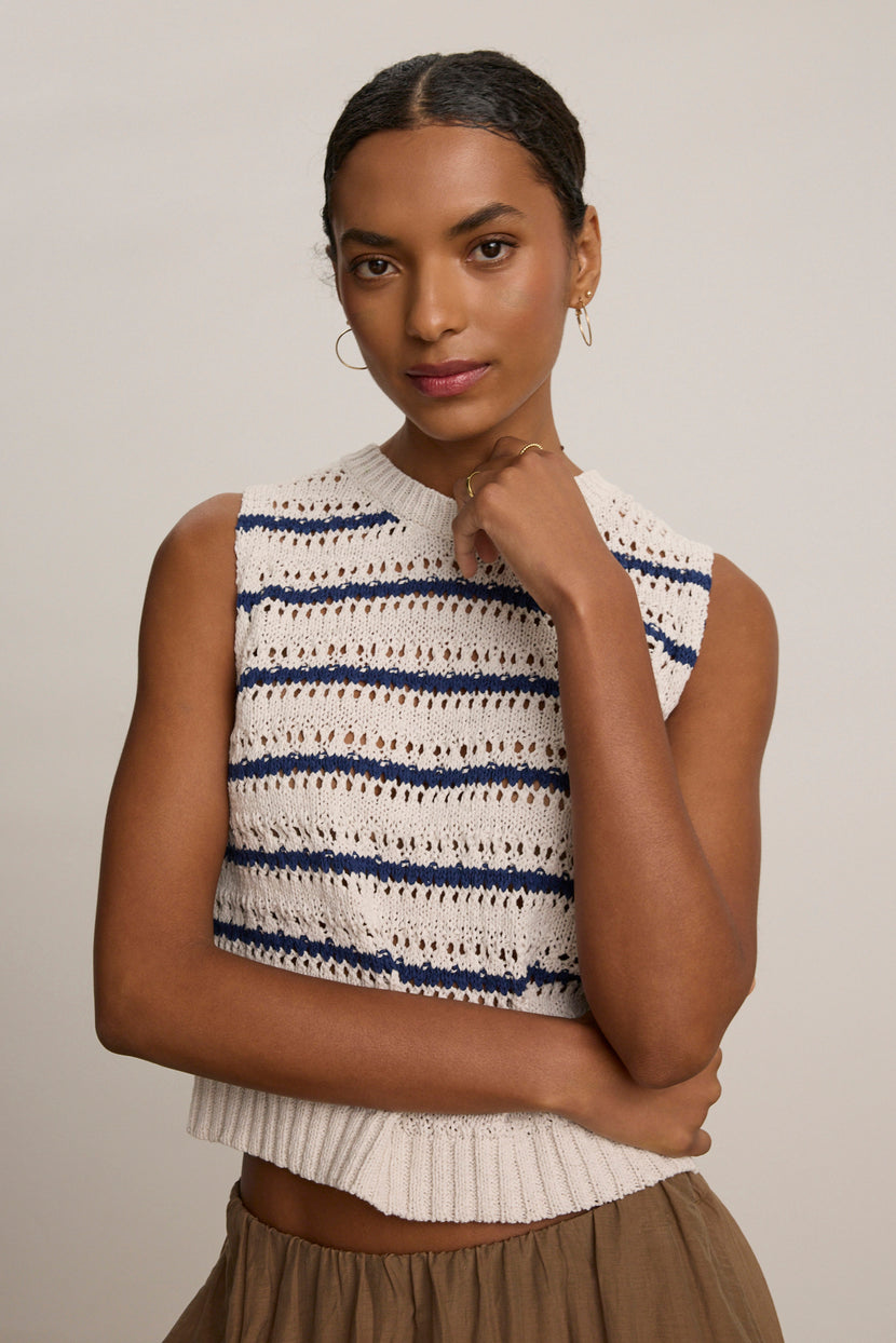 A woman stands with arms crossed, wearing the CALISTA SWEATER VEST by Velvet by Graham & Spencer—a blue and white striped sleeveless knit cropped vest—paired with tan pants, looking at the camera against a plain background.
