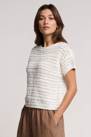 A woman faces forward against a plain background, wearing the cream-colored BLOSSOM SWEATER by Velvet by Graham & Spencer with short sleeves and lightweight fabric, paired with light brown pants.