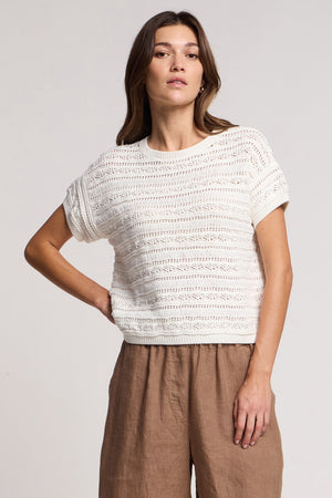 A woman poses against a plain background wearing the BLOSSOM SWEATER by Velvet by Graham & Spencer—a white, lightweight summer knit—paired with high-waisted brown pants, one hand resting on her hip.