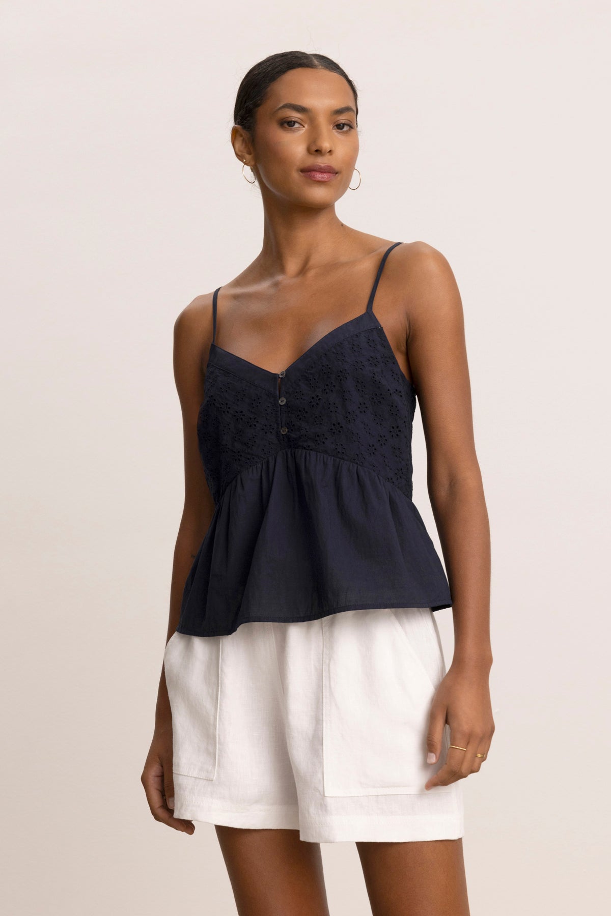 A woman wears the ALIAH EYELET TANK TOP by Velvet by Graham & Spencer—a navy blue, sleeveless, tiered top with thin straps—paired with white shorts, standing against a plain beige background.-39123260571841