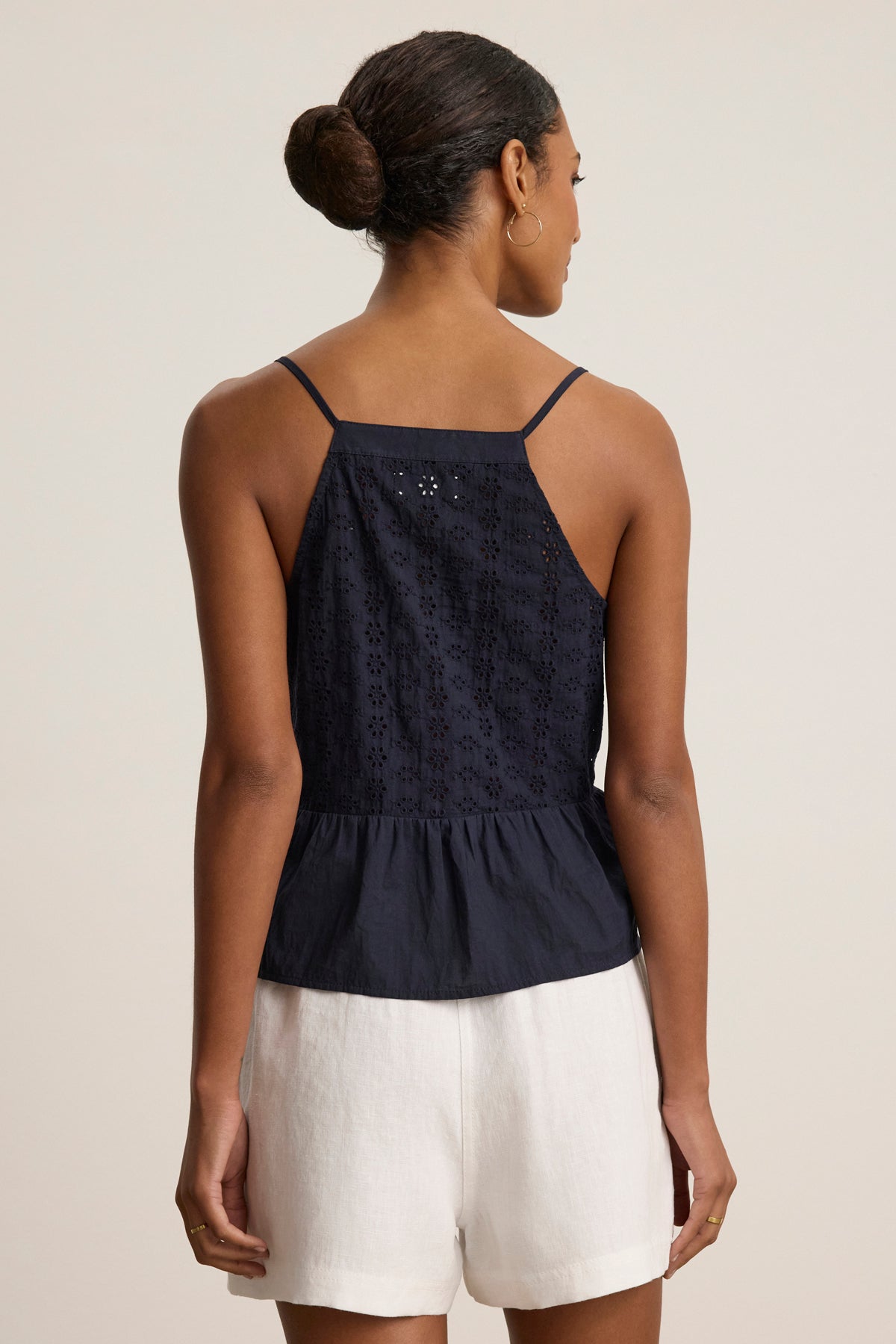 A woman stands with her back to the camera, wearing the ALIAH EYELET TANK TOP by Velvet by Graham & Spencer in navy blue, paired with white shorts against a plain light background.-39123260735681