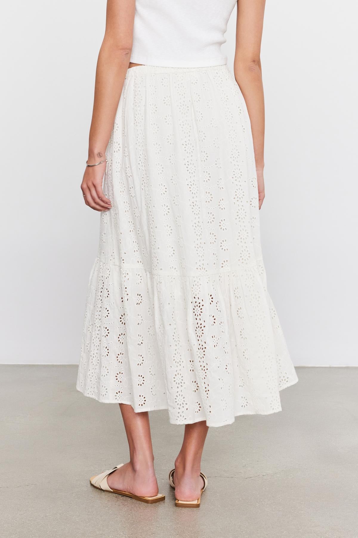   Woman wearing a white, cotton broderie anglaise Amelia skirt by Velvet by Graham & Spencer with eyelet details, paired with a simple top and gold flat shoes, standing against a neutral background. 