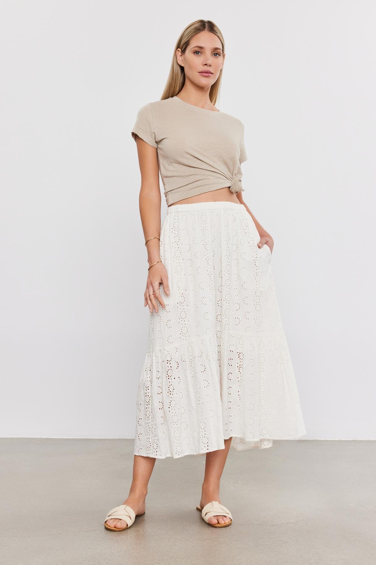   A woman stands in a studio, wearing a beige crop top, an AMELIA SKIRT from Velvet by Graham & Spencer with eyelet details, and white sandals. 