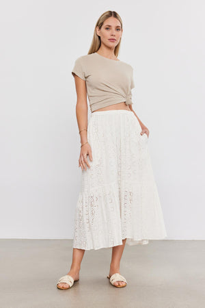 A woman stands in a studio, wearing a beige crop top, an AMELIA SKIRT from Velvet by Graham & Spencer with eyelet details, and white sandals.