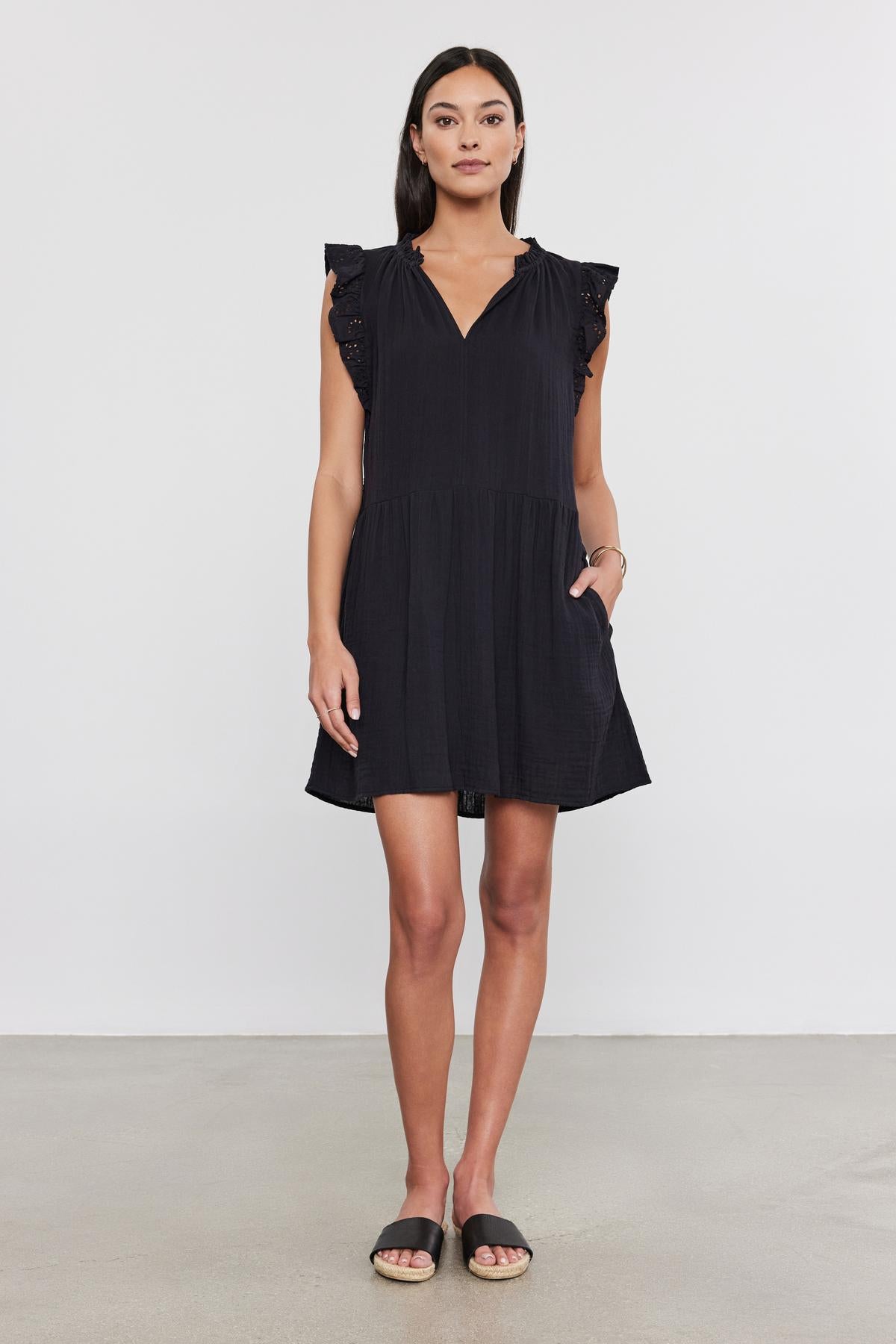   Woman in a black cotton gauze Grace dress by Velvet by Graham & Spencer with flutter sleeves, standing indoors, facing the camera with a neutral expression. She wears simple black sandals. 