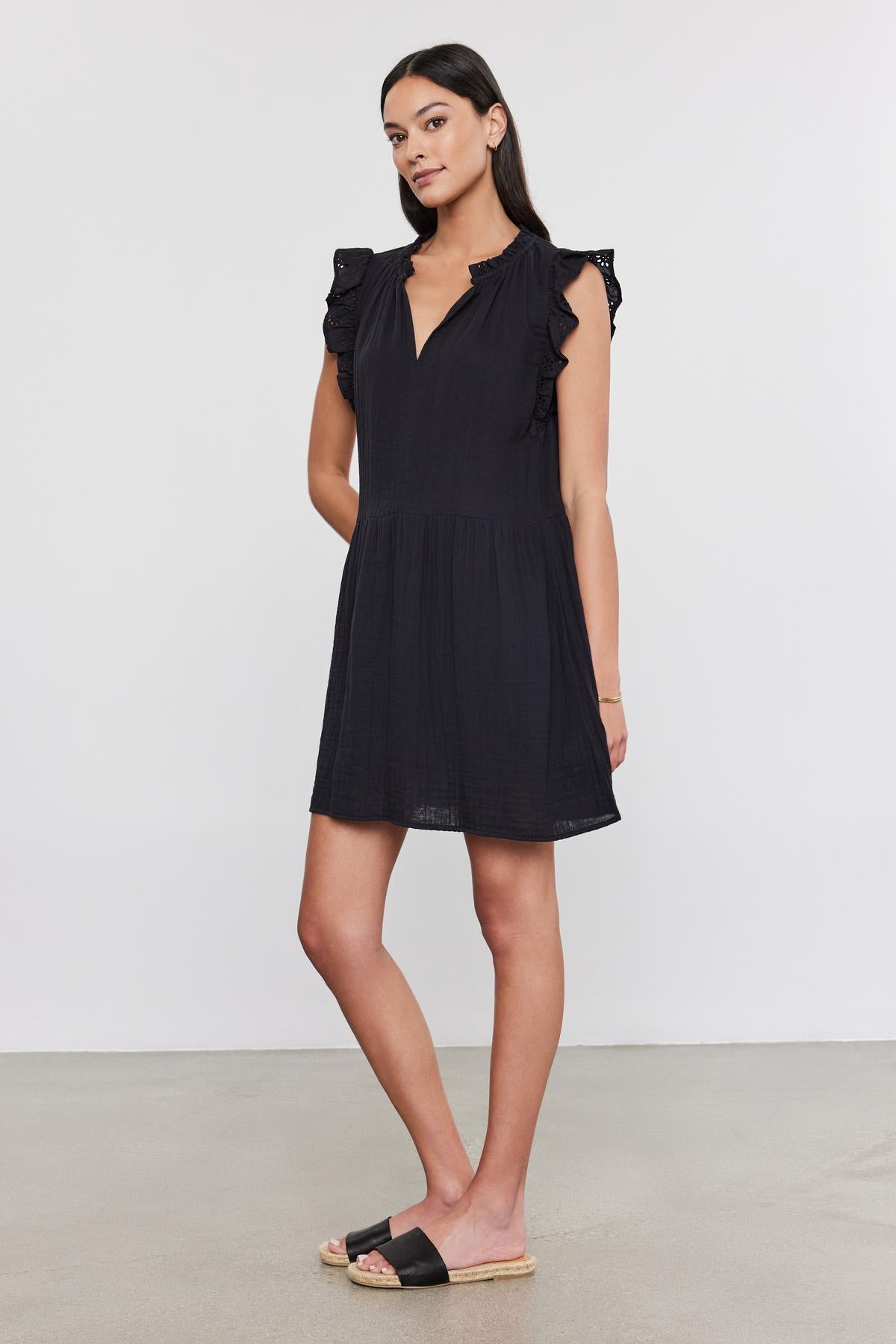   A woman in a Velvet by Graham & Spencer GRACE DRESS with cotton eyelet flutter sleeves, standing against a plain background, wearing black and beige sandals. 