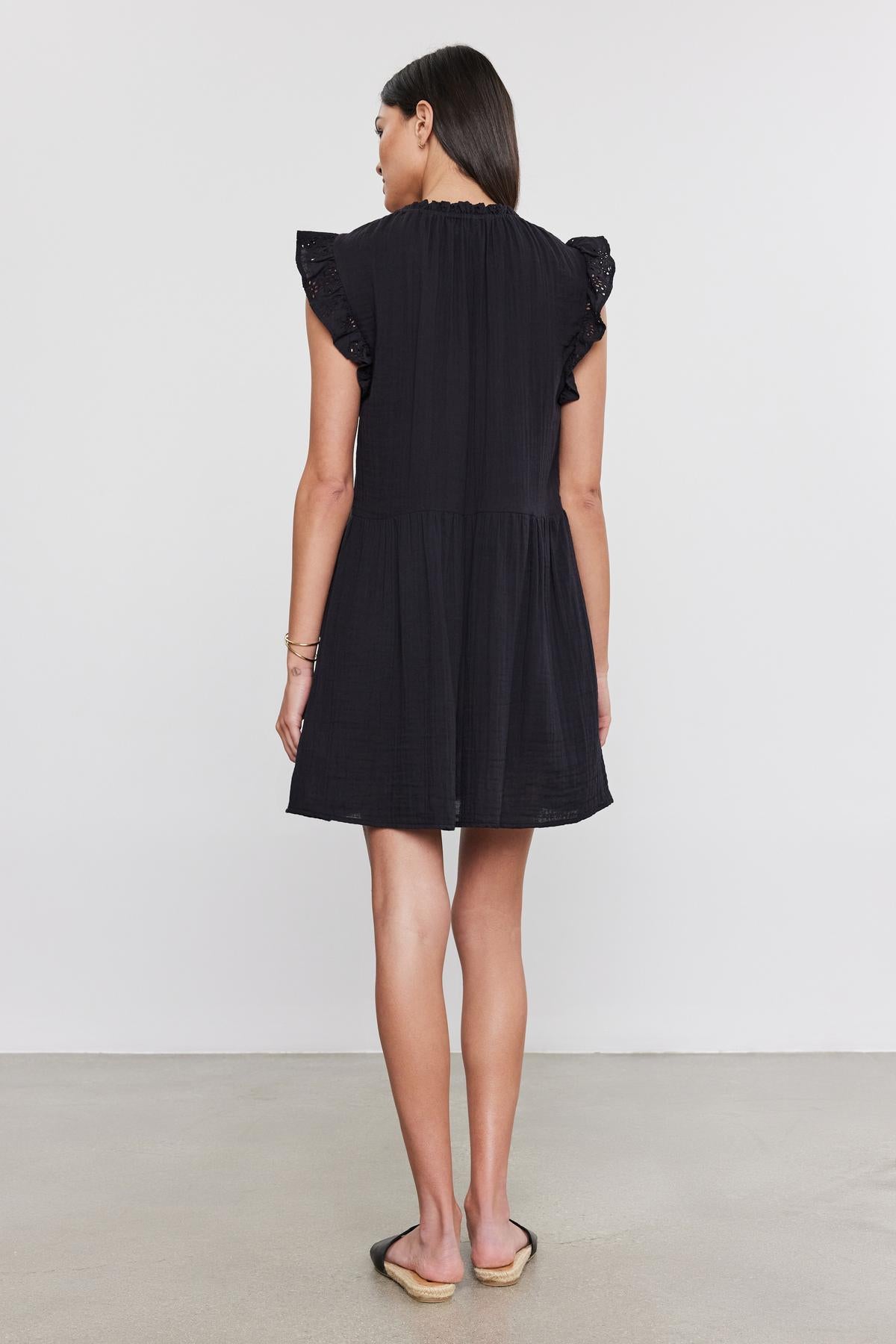 A woman in a black Velvet by Graham & Spencer dress with cotton eyelet flutter sleeves stands facing away, on a plain light background.-36910266253505