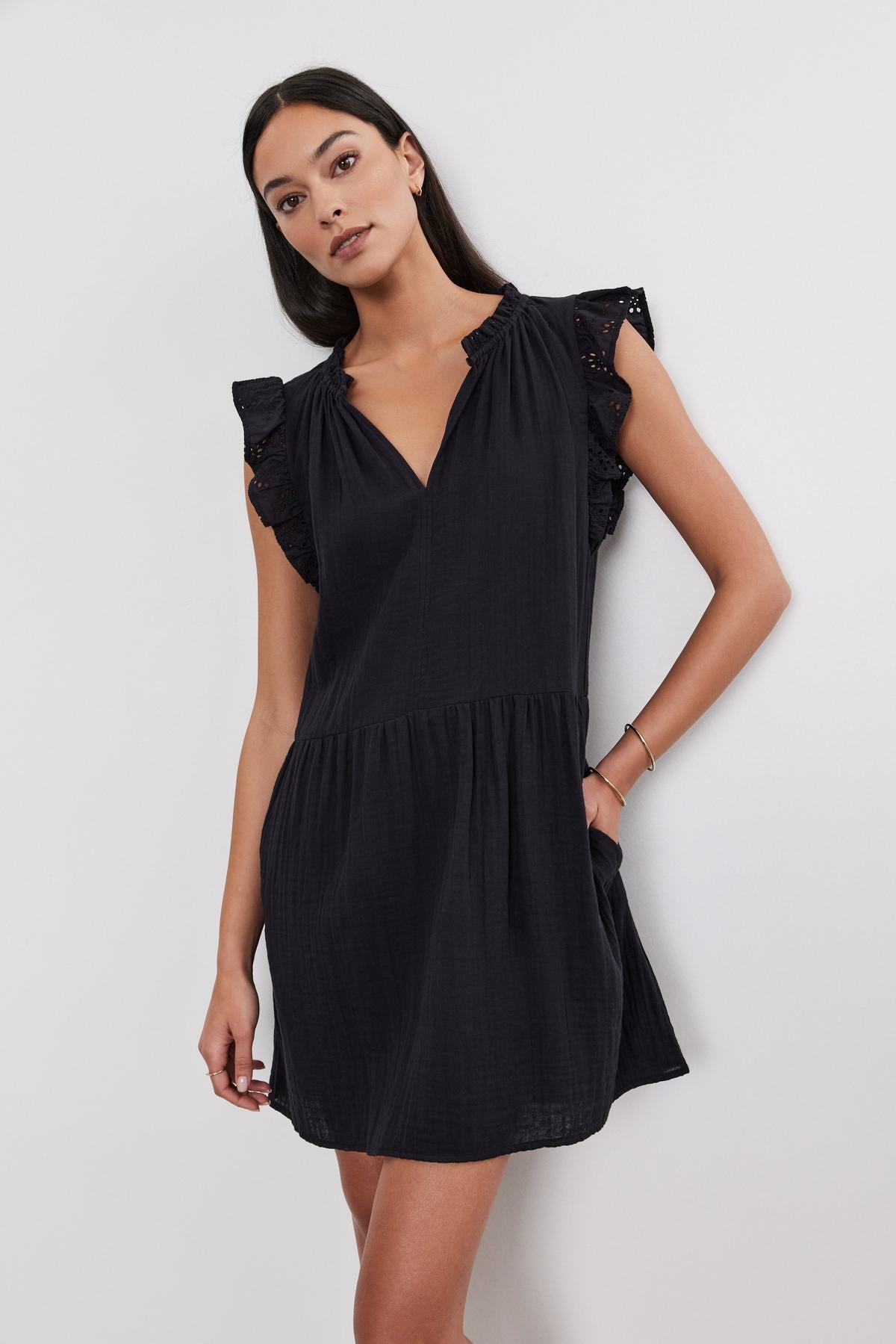   A woman in a black, cotton eyelet flutter sleeve Grace dress by Velvet by Graham & Spencer stands against a white background, looking to her left with a neutral expression. 