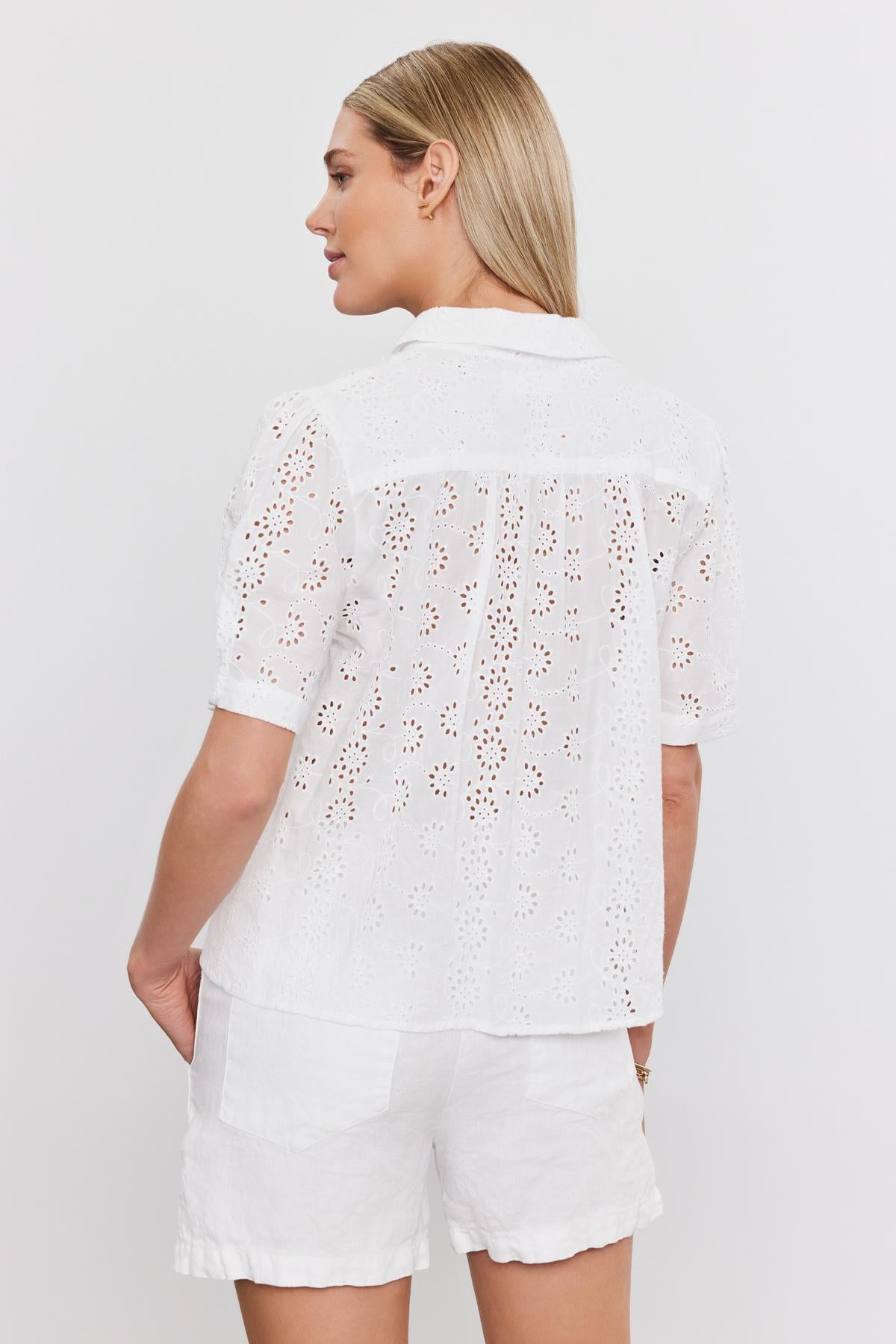   A woman from behind wearing a white, short-sleeved, cotton Olivia blouse with eyelet details and matching shorts, standing against a plain background by Velvet by Graham & Spencer. 