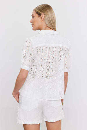 A woman from behind wearing a white, short-sleeved, cotton Olivia blouse with eyelet details and matching shorts, standing against a plain background by Velvet by Graham & Spencer.