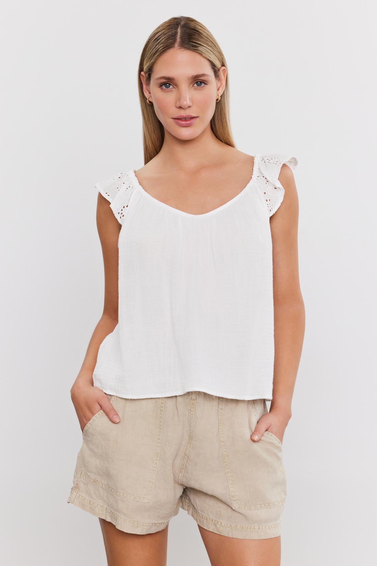   A person wearing the STELLA TANK TOP by Velvet by Graham & Spencer, which is a white sleeveless top with ruffled straps, along with beige shorts, posing with hands in pockets against a plain white background. 