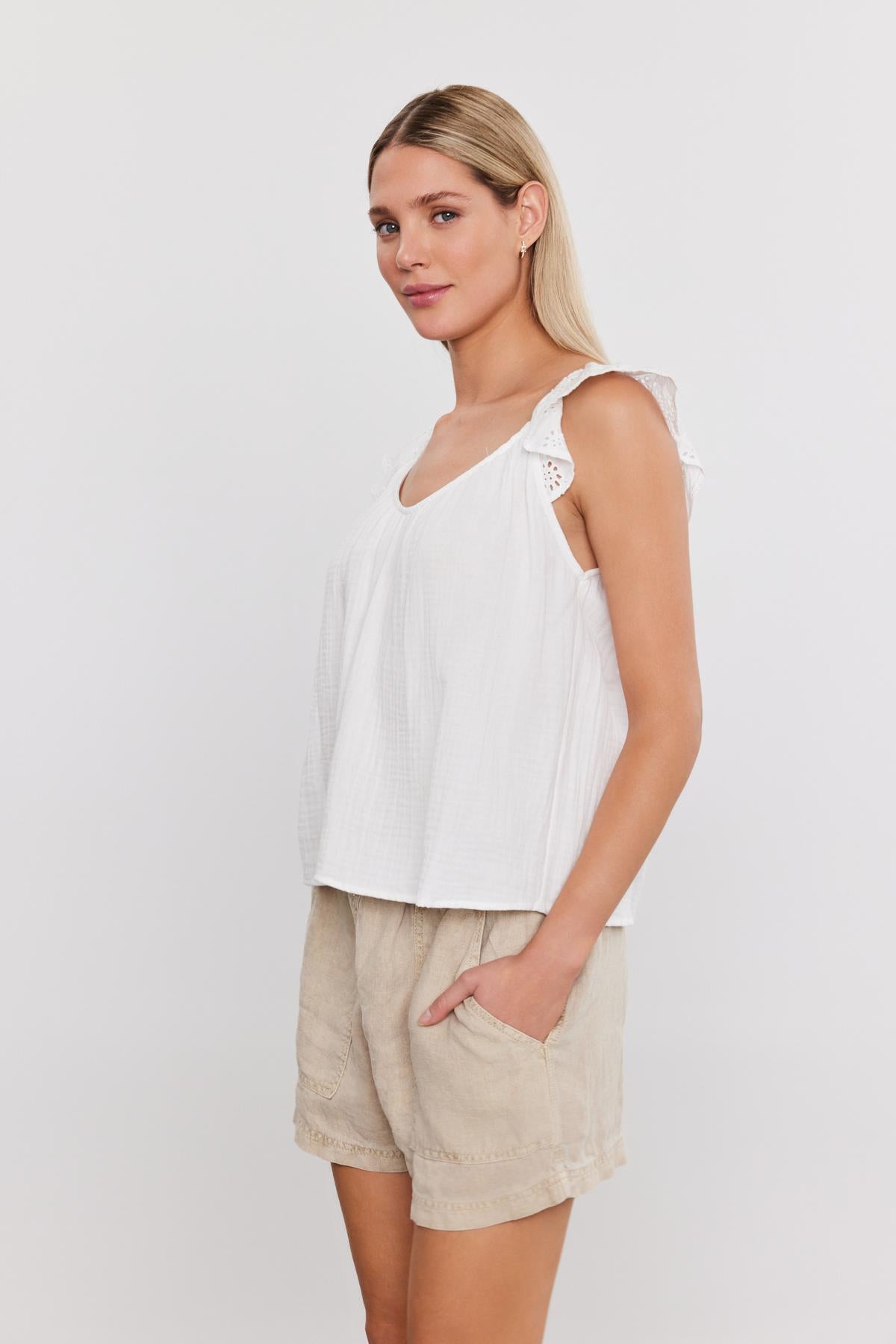   A person stands facing slightly left, wearing the white STELLA TANK TOP by Velvet by Graham & Spencer, which features ruffled shoulders, paired with beige shorts. 