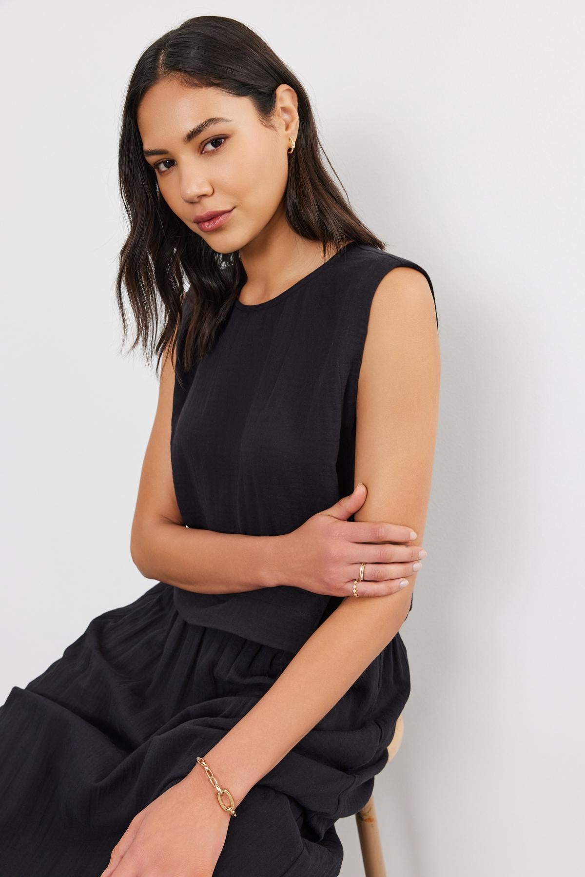   A woman in a black Velvet by Graham & Spencer AUBREN COTTON GAUZE TANK TOP and skirt, sitting and looking at the camera with a subtle smile. 