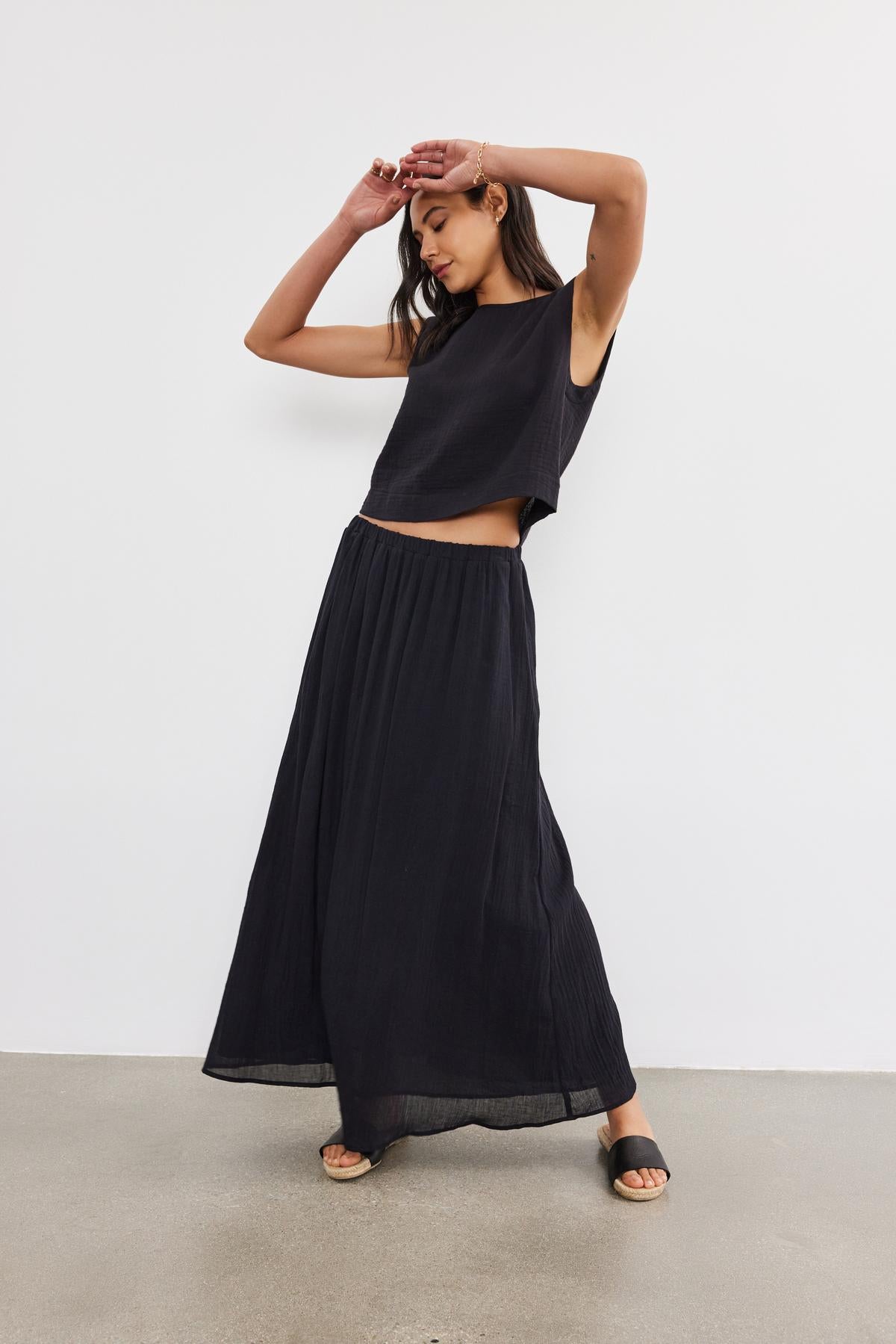   A woman in a black crop top and the Velvet by Graham & Spencer INDY COTTON GAUZE SKIRT poses in a studio, one hand raised to her forehead, looking downwards. 