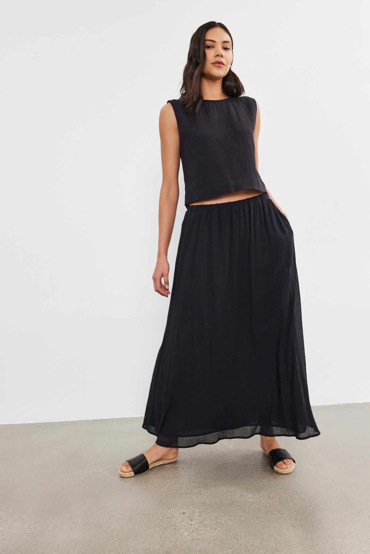 A woman wearing a black Aubren Cotton Gauze tank top and a long matching skirt stands on a plain background, looking at the camera.-36910092091585