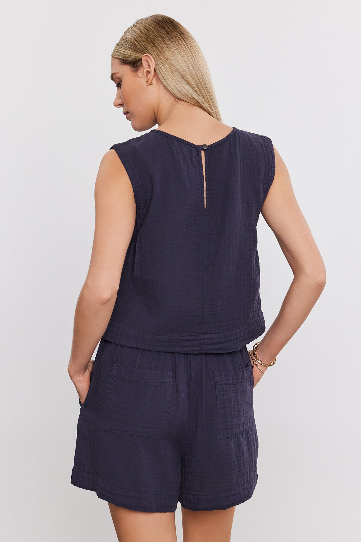   Woman wearing the AUBREN COTTON GAUZE TANK TOP by Velvet by Graham & Spencer, a sleeveless, navy blue top and matching shorts, viewed from the back. Made of airy cotton gauze, the ensemble features a cropped silhouette that adds to its casual charm. She has her hands in her pockets. 