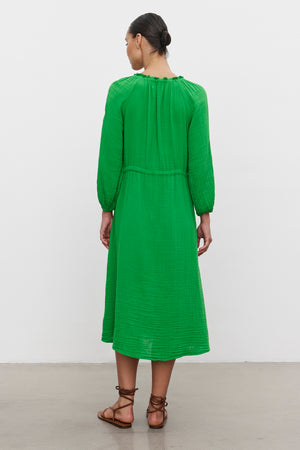 Woman standing with her back to the camera, wearing a green mid-length warm weather AUDREY COTTON GAUZE DRESS with gathered detailing, accessorized with brown sandals by Velvet by Graham & Spencer.