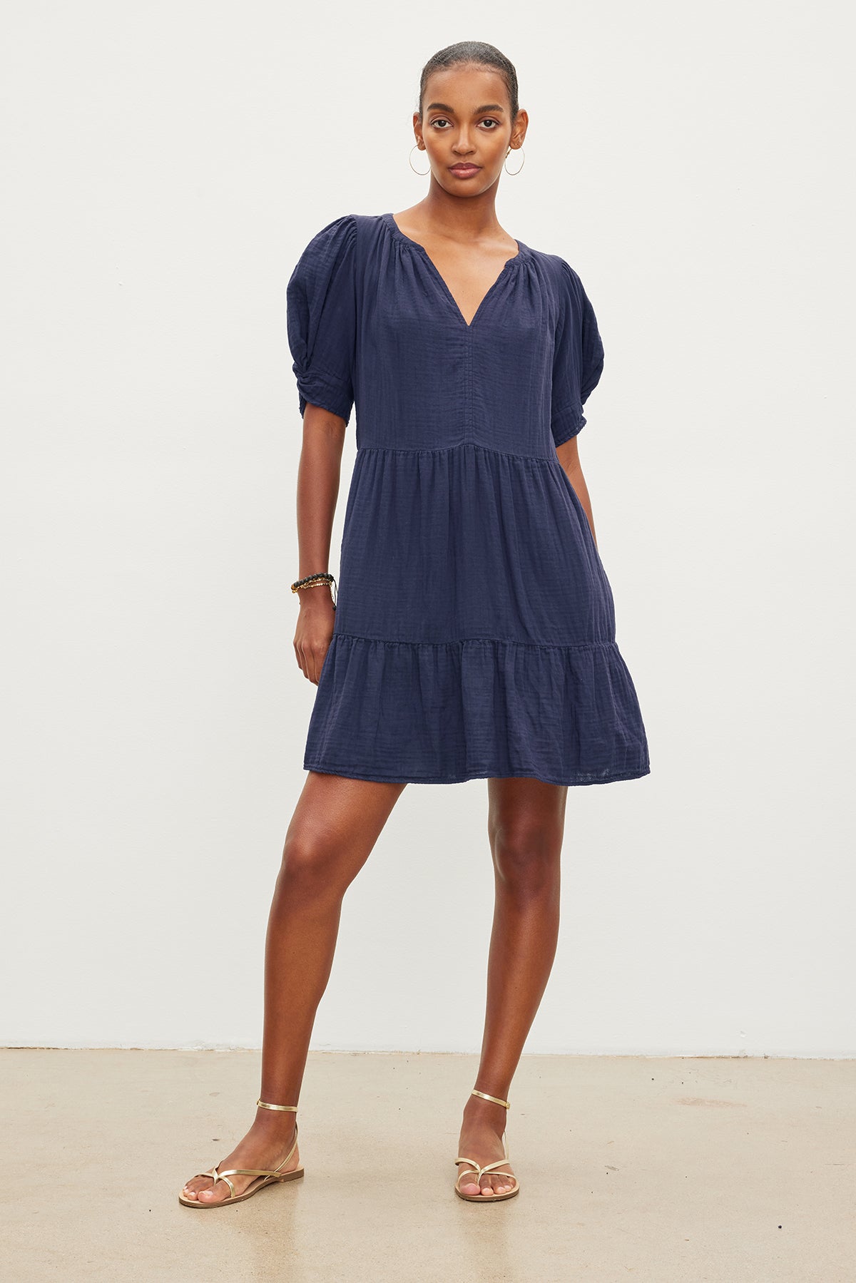 A person stands against a plain white background, wearing a navy blue knee-length BELLA COTTON GAUZE DRESS with short puff sleeves and gold sandals. The v-neckline dress by Velvet by Graham & Spencer features a detachable belt, adding versatility. They are looking directly at the camera with one hand on their hip.-37355941626049