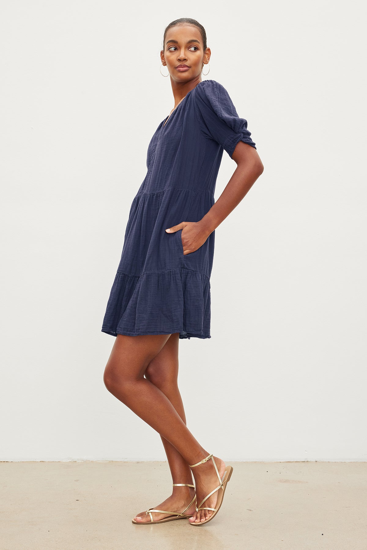   A person stands sideways against a plain background, wearing a navy blue BELLA COTTON GAUZE DRESS by Velvet by Graham & Spencer and gold sandals. 