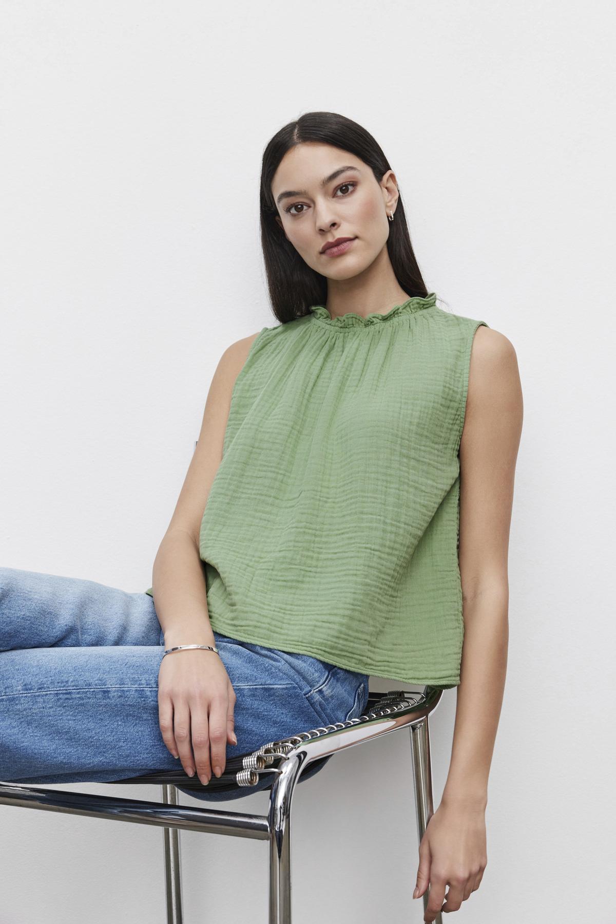   Woman in a green Velvet by Graham & Spencer BIANCA COTTON GAUZE TANK TOP with an elastic neckline and blue jeans sitting on a chrome chair against a white background. 