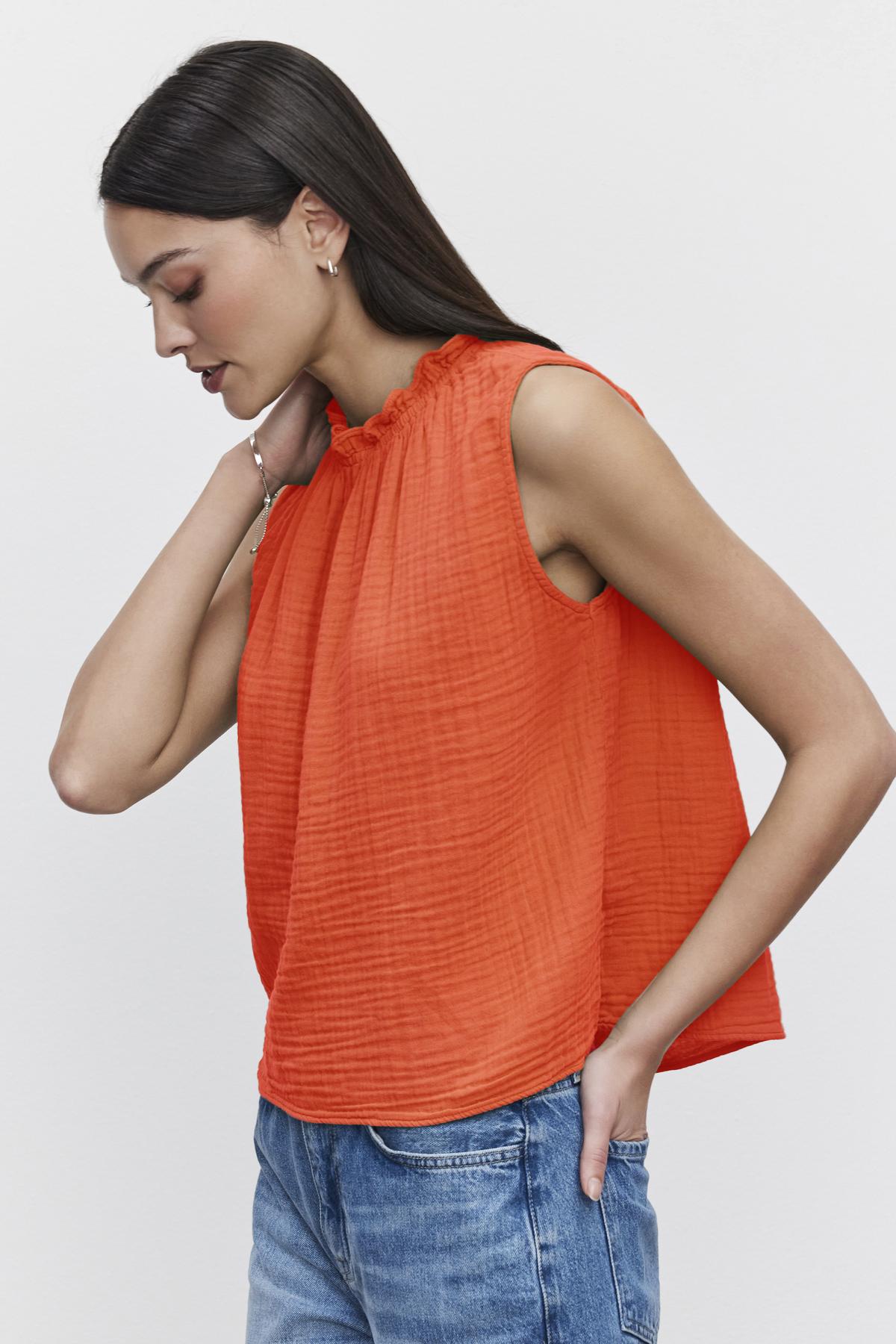   A woman with long dark hair is wearing a sleeveless, orange, ruffled BIANCA COTTON GAUZE TANK TOP by Velvet by Graham & Spencer and blue jeans, standing against a plain white background. She is looking down with her right hand touching her neck and left hand in her pocket. 