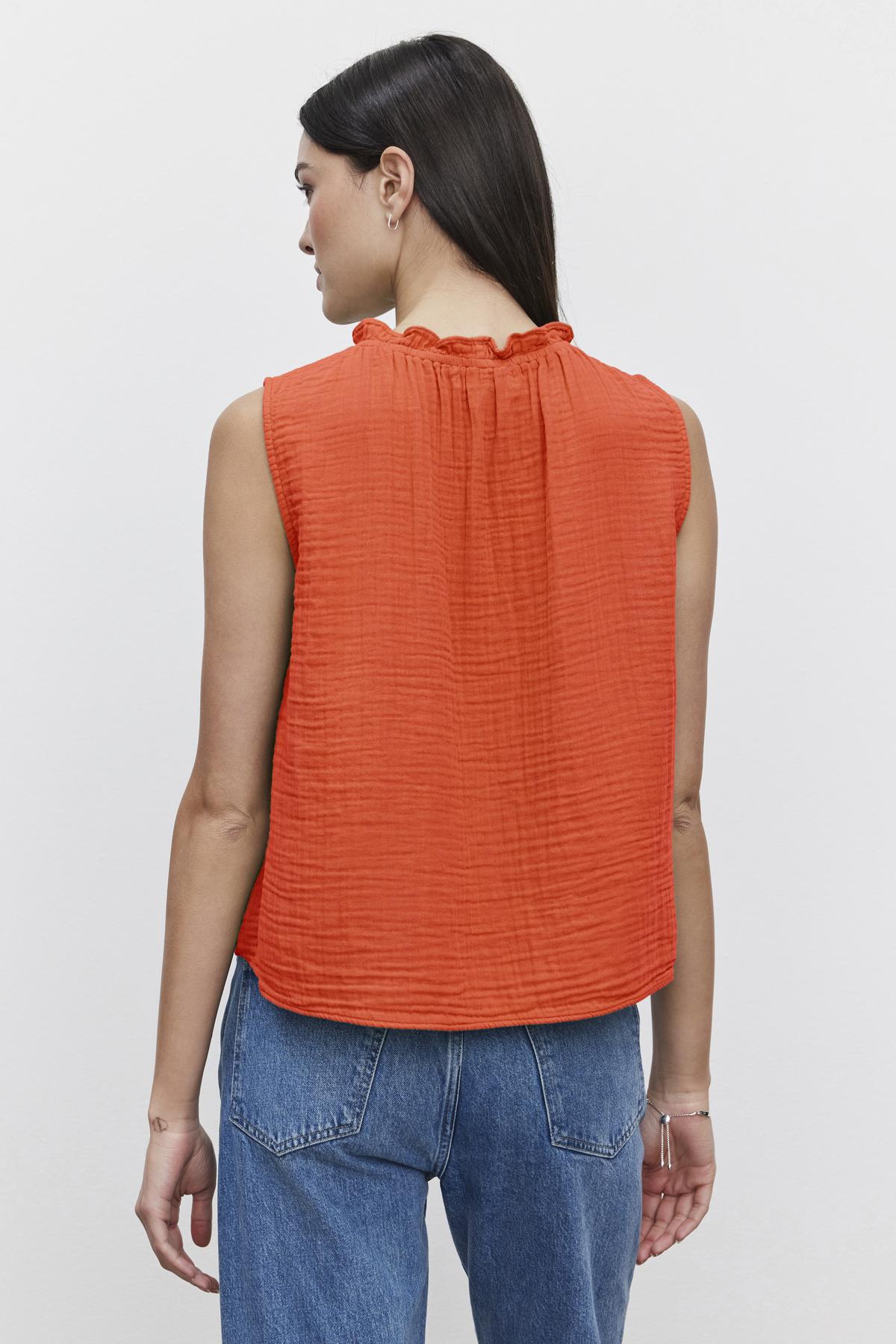   A woman with long straight hair is wearing a sleeveless orange BIANCA COTTON GAUZE TANK TOP by Velvet by Graham & Spencer and blue jeans. She is facing away from the camera, showing the back of her outfit. 