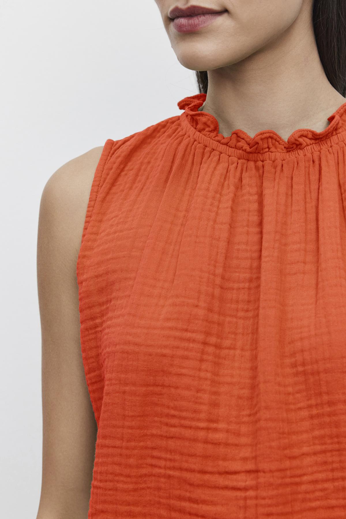   Close-up of a person wearing an orange, sleeveless warm weather blouse made from textured cotton gauze, featuring a ruffled, elastic neckline. The background is plain white. Only the shoulders, neck, and lower half of the face are visible. The blouse is identified as the 