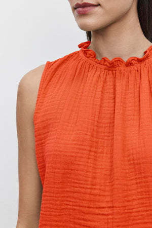 Close-up of a person wearing an orange, sleeveless warm weather blouse made from textured cotton gauze, featuring a ruffled, elastic neckline. The background is plain white. Only the shoulders, neck, and lower half of the face are visible. The blouse is identified as the 