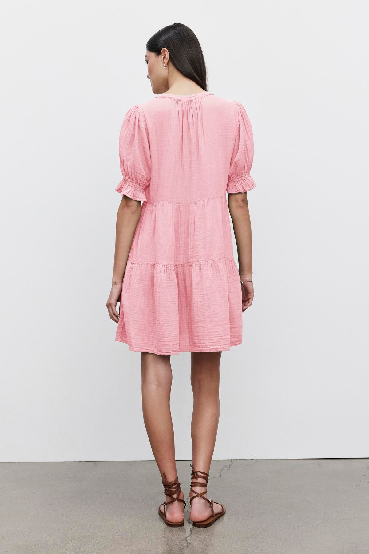   A person with long hair is standing facing away, wearing the CLARISSA COTTON GAUZE TIERED DRESS by Velvet by Graham & Spencer and brown lace-up sandals. 