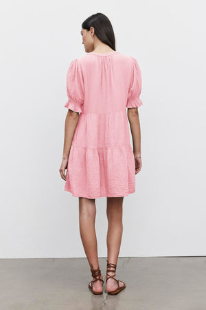 A person with long hair is standing facing away, wearing the CLARISSA COTTON GAUZE TIERED DRESS by Velvet by Graham & Spencer and brown lace-up sandals.